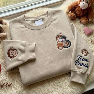 Puede incluir: Sudadera beige con diseño de mono bordado. Presenta un gráfico de mono en la manga izquierda y un diseño más grande de dos monos abrazándose en el pecho. La manga derecha tiene el texto "Team Punch" en azul.