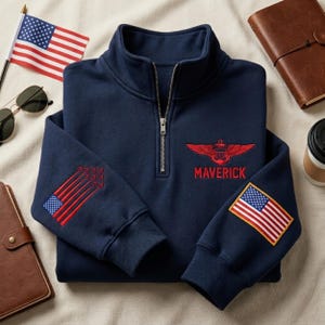 May include: Navy blue quarter-zip sweatshirt with embroidered red wings and the word "MAVERICK." Features an American flag patch on the sleeve and a smaller flag design on the other sleeve. Includes a small American flag and sunglasses.