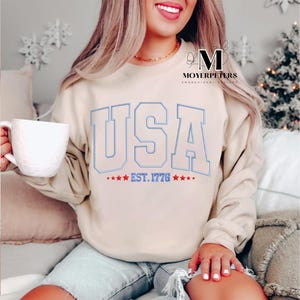 May include: A beige sweatshirt with a white graphic of "USA EST. 1776" with blue outlines and red stars.