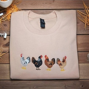 May include: A light tan sweatshirt featuring an embroidered design of four chickens in a row. The chickens are in various colors, including white, black, brown, and red, with yellow feet. The sweatshirt is made of a soft material.