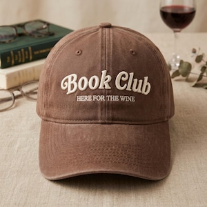 Może przedstawiać: Brązowa czapka z daszkiem z wyhaftowanym białym napisem Book Club oraz mniejszym tekstem HERE FOR THE WINE wyśrodkowanym na przednim panelu. Czapka posiada usztywnianą koronę, zakrzywiony daszek oraz przeszywane otwory wentylacyjne.