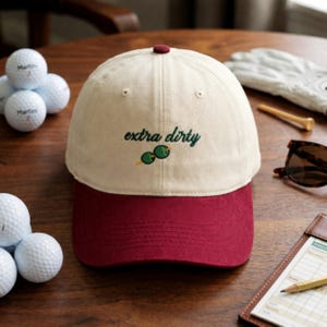Puede incluir: Gorra de b&eacute;isbol crema y burdeos con la frase bordada "extra dirty" y un dise&ntilde;o de aceitunas. La gorra se muestra con pelotas de golf y accesorios. El dise&ntilde;o de la gorra es adecuado para un entusiasta del golf.