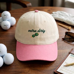 Puede incluir: Gorra de b&eacute;isbol beige y rosa con el texto bordado "extra dirty" en verde. La gorra tiene una visera rosa y un bot&oacute;n rosa en la parte superior. Se ven pelotas de golf y gafas de sol en el fondo.