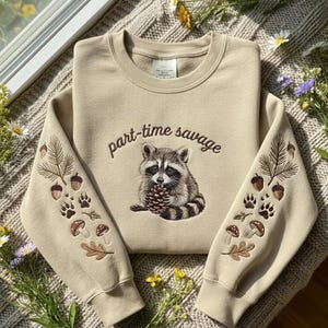 Puede incluir: Sudadera beige con un mapache sosteniendo una piña, el texto "part-time savage", y detalles bordados de bellotas, huellas de patas, setas y hojas en las mangas. La sudadera está colocada sobre una superficie texturizada.