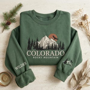 Könnte beinhalten: Grünes Sweatshirt mit gestickter Colorado-Berglandschaft, mit einer Bergkette, Bäumen und einer untergehenden Sonne. Das Wort "COLORADO" ist unterhalb der Berglandschaft gestickt, darunter "ROCKY MOUNTAIN". Auf dem linken Ärmel steht "WILDER".