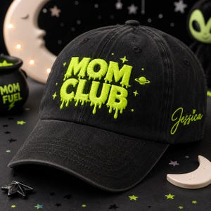 Op de afbeelding: Een zwarte baseballpet met neon-groen geborduurde tekst die MOM CLUB leest in een druipend lettertype. Het ontwerp bevat kleine ster-iconen en een planeet-afbeelding. De naam Jessica is in neon-groen cursief geborduurd aan de zijkant.