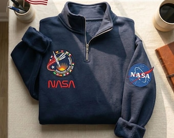 May include: Navy blue sweatshirt with a quarter-zip collar and NASA patches. The sweatshirt features a NASA logo patch on the sleeve and a colorful mission patch on the chest, with the NASA name in red.