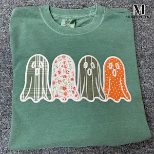 Può includere: Una maglietta verde girocollo con quattro applicazioni a forma di fantasma. Ogni fantasma ha un motivo diverso: a quadri, floreale, a righe e a pois. La maglietta è piegata su una superficie strutturata.