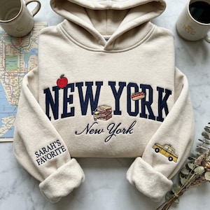以下が含まれることがあります： クリーム色のフード付きスウェットシャツ。ネイビーブルーの刺繍で「NEW YORK」とリンゴ、サンドイッチ、ホットドッグのグラフィック。袖には「SARAH'S FAVORITE」と黄色のタクシーが描かれています。