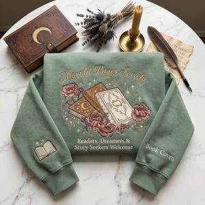 May include: Sage green sweatshirt with embroidered design. The design includes the text "Moonlit Pages Society", books, flowers, and stars. The sleeves have an embroidered open book and the words "Book Coven". The text "Readers, Dreamers & Story Seekers Welcome" is also included.