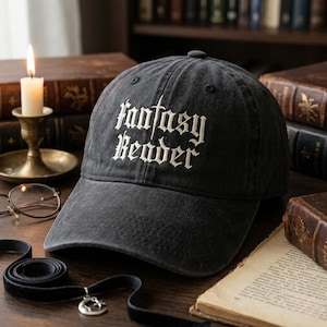 May include: A dark grey baseball cap featuring the embroidered words "Fantasy Reader" in white gothic script. The cap has a slightly worn appearance and a curved peak. The background includes books, a candle, and spectacles.