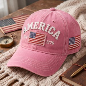 May include: A pink baseball cap featuring the white embroidered text AMERICA, the text EST 1776, and a rectangular American flag patch on the front. A smaller American flag is embroidered on the side of the cap, which has white stitching.