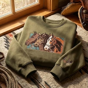 Op de afbeelding: Olijfgroene sweatshirt met een rechthoekige afbeelding van drie paarden met gedetailleerde hoofdstellen. De mouw heeft een geborduurd hoefijzer en een veermotief. Het kledingstuk ligt op een patroonkleed.