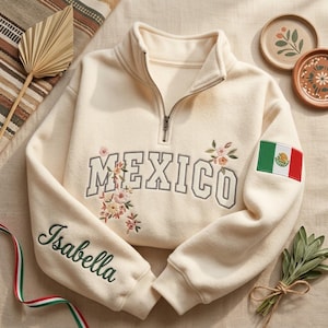 May include: Cream-colored sweatshirt with a quarter-zip neckline. The word "MEXICO" is embroidered across the chest with floral accents. The sleeve features the name "Isabella" and the Mexican flag. A stylish and personalized garment.