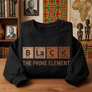May include: Black crewneck sweatshirt with the word "BLACK" spelled out using elements from the periodic table. The text "THE PRIME ELEMENT" is below the word. The sweatshirt is displayed on a wooden surface.