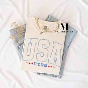 May include: A cream colored t-shirt with a blue and white embroidered "USA" graphic and the text "EST. 1776" with red stars.