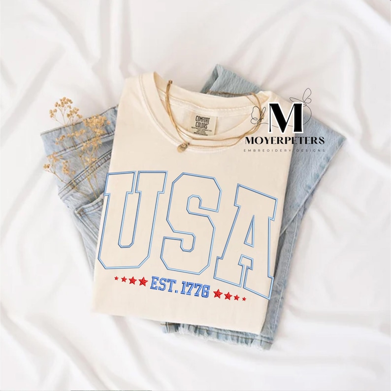 May include: A cream colored t-shirt with a blue and white embroidered "USA" graphic and the text "EST. 1776" with red stars.