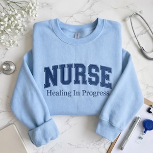 Puede incluir: Sudadera azul claro con la palabra "NURSE" en letras de bloque azul marino en el pecho. Debajo, la frase "Healing In Progress" está impresa en azul marino. Un estetoscopio y otros accesorios médicos son visibles.