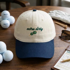 Puede incluir: Gorra de b&eacute;isbol crema y azul marino con la inscripci&oacute;n bordada "extra dirty" en verde y un gr&aacute;fico de dos aceitunas. Se ven pelotas de golf, gafas de sol y una tarjeta de puntuaci&oacute;n.