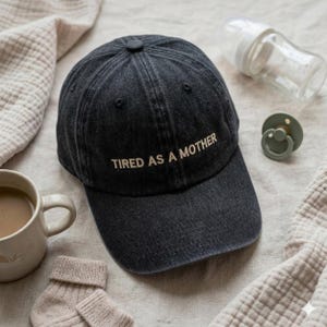 Peut inclure: Une casquette de baseball en jean délavé gris anthracite avec le texte brodé TIRED AS A MOTHER en beige clair. La casquette présente une calotte structurée, une visière incurvée et un bouton sur le dessus, sur un fond aux tons neutres.