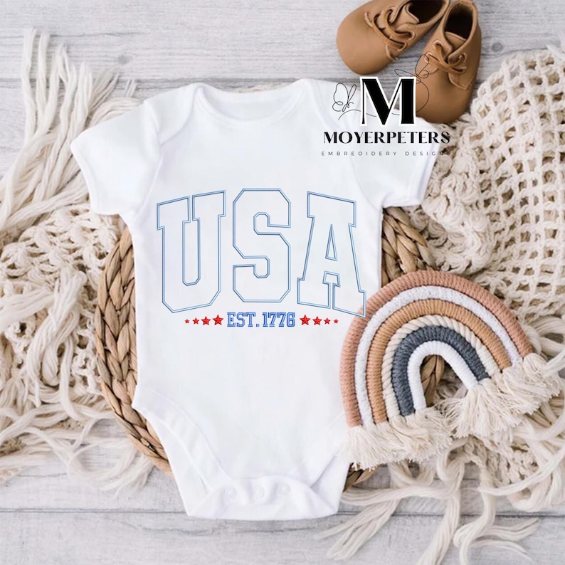 May include: White baby onesie with blue and red embroidery that says "USA EST. 1776" with stars.
