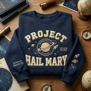 May include: Navy blue sweatshirt with the words "PROJECT INTERSTELLAR MISSION HAIL MARY" in a cream-colored, textured font. The design includes a planet with rings, a rocket, and stars. The sleeve has a rocket, planet, and star design.