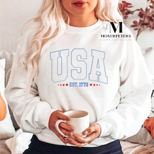 May include: White sweatshirt with a blue outline of the word "USA" and the text "EST. 1776" in red.