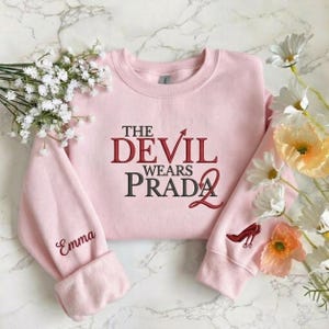 May include: Pale pink sweatshirt featuring the text "THE DEVIL WEARS PRADA 2" in burgundy and black. The left sleeve has the name "Emma" embroidered in burgundy, and the right sleeve has a red high-heeled shoe graphic.
