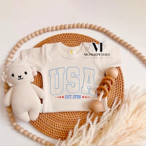 May include: A white baby onesie with blue and red embroidery that says "USA EST. 1776" with stars. The onesie is on a woven wicker surface with a wooden rattle and a knitted white lamb toy.