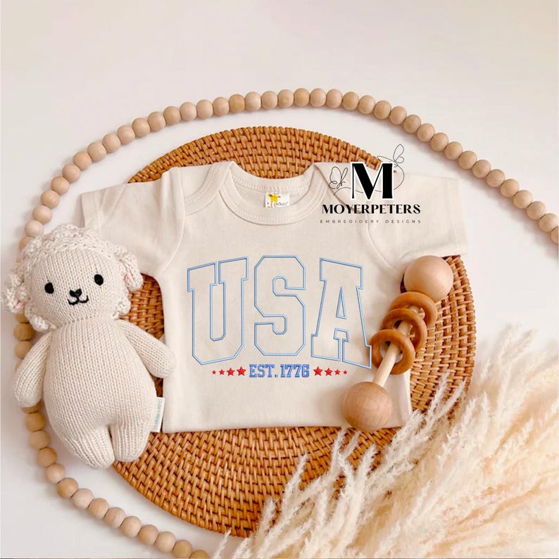 May include: A white baby onesie with blue and red embroidery that says "USA EST. 1776" with stars. The onesie is on a woven wicker surface with a wooden rattle and a knitted white lamb toy.
