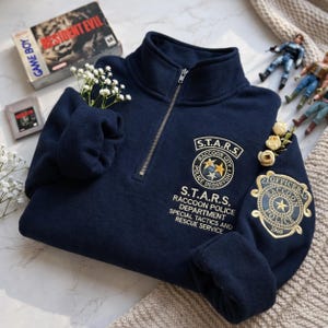 May include: Navy blue zip-up sweatshirt featuring embroidered patches with the text "S.T.A.R.S. Raccoon Police Department." The sweatshirt has a high collar and a silver zipper. The design includes the text "Resident Evil" on a Game Boy box.