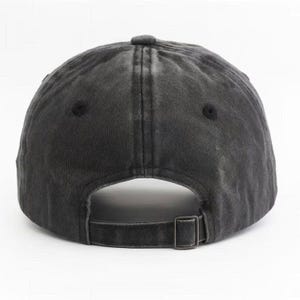 May include: A charcoal grey baseball cap with a washed, distressed finish. The cap features a six-panel construction with stitched eyelets and an adjustable fabric strap at the back, secured with a rectangular metal buckle for a custom fit.