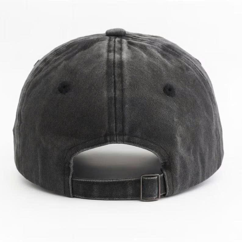 May include: A charcoal grey baseball cap with a washed, distressed finish. The cap features a six-panel construction with stitched eyelets and an adjustable fabric strap at the back, secured with a rectangular metal buckle for a custom fit.