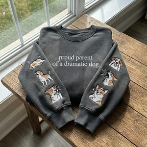May include: Dark gray sweatshirt with the phrase "proud parent of a dramatic dog" in white script. The sleeves feature embroidered images of dogs in various dramatic poses. The sweatshirt is displayed on a wooden surface.