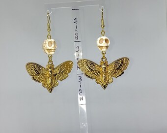 May include: A pair of gold-colored earrings with a unique design. Each earring features a small, off-white skull bead, a gold-colored moth pendant, and a gold-colored hook. The moth pendants are approximately 1.5 inches wide.