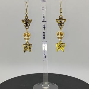 May include: Gold tone earrings with a white skull bead, a gold butterfly charm, and a smaller butterfly charm with rhinestones. The earrings are displayed on a clear acrylic stand with a ruler that reads "1 inch, 2 inch, 3 inch".