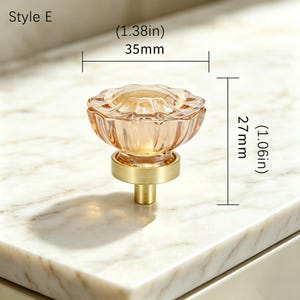 May include: A Style E cabinet knob featuring a translucent amber-colored glass top with a scalloped edge and a brushed brass base. The knob measures 1.38 inches in diameter and 1.06 inches in height, as indicated by the technical dimension labels.