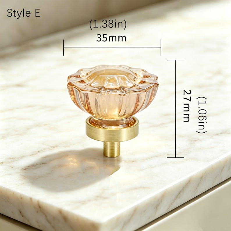 May include: A Style E cabinet knob featuring a translucent amber-colored glass top with a scalloped edge and a brushed brass base. The knob measures 1.38 inches in diameter and 1.06 inches in height, as indicated by the technical dimension labels.