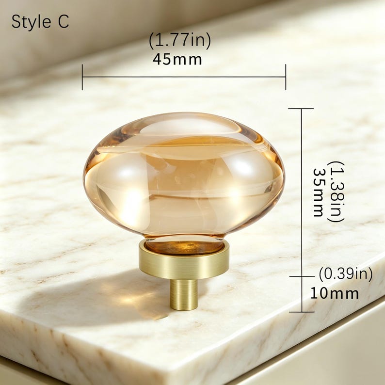 May include: Style C amber-colored glass knob with a brushed gold metal base. The knob measures 1.77 inches in width, 1.38 inches in total height, and the base stem measures 0.39 inches in height. The design features a smooth, rounded shape.