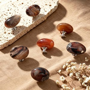 May include: A collection of six oval-shaped cabinet knobs featuring polished, multi-colored agate stone tops with swirling patterns in shades of brown, tan, and cream, each mounted on a small, circular silver-toned metal base.