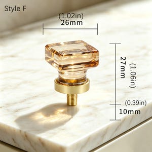 May include: A square amber-colored glass cabinet knob with a brushed gold metal base. The knob features a threaded metal screw at the bottom. Dimensions are labeled as 1.02 inches wide, 1.06 inches high, and a 0.39 inch screw length. Text reads Style F.