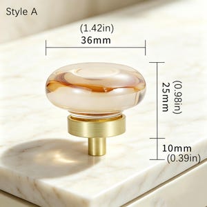 May include: A round cabinet knob labeled Style A, featuring a translucent amber glass top and a brushed gold metal base. The knob measures 1.42 inches (36mm) in width, 0.98 inches (25mm) in height, and has a 0.39 inch (10mm) base height.