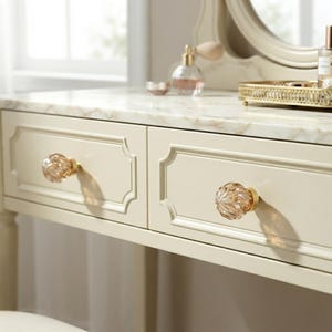May include: A cream-colored vanity table featuring a white marble top with grey veining. The table has two drawers, each equipped with a round, faceted, clear glass knob with a gold-toned metal base. A gold-toned tray and perfume bottle sit on top.