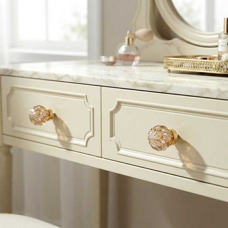 May include: A cream-colored vanity table featuring a white marble top with grey veining. The table has two drawers, each equipped with a round, faceted, clear glass knob with a gold-toned metal base. A gold-toned tray and perfume bottle sit on top.