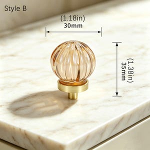 May include: A round, amber-colored glass cabinet knob with a ribbed texture, mounted on a brushed gold-tone metal base. The knob is labeled Style B and features dimensions of 1.18 inches in width and 1.38 inches in total height.