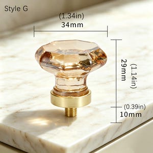May include: Style G cabinet knob with a faceted, light amber-colored glass top and a brushed gold-tone metal base. The knob measures 1.34 inches wide, 1.14 inches high, and has a 0.39 inch threaded post for installation on furniture or cabinetry.