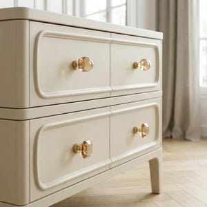 May include: A cream-colored chest of drawers featuring four rectangular drawer fronts with rounded edges. Each drawer is equipped with a single, faceted, translucent amber-toned glass knob, providing a clear contrast to the solid light-colored frame.