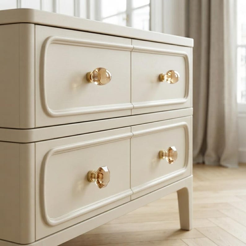 May include: A cream-colored chest of drawers featuring four rectangular drawer fronts with rounded edges. Each drawer is equipped with a single, faceted, translucent amber-toned glass knob, providing a clear contrast to the solid light-colored frame.