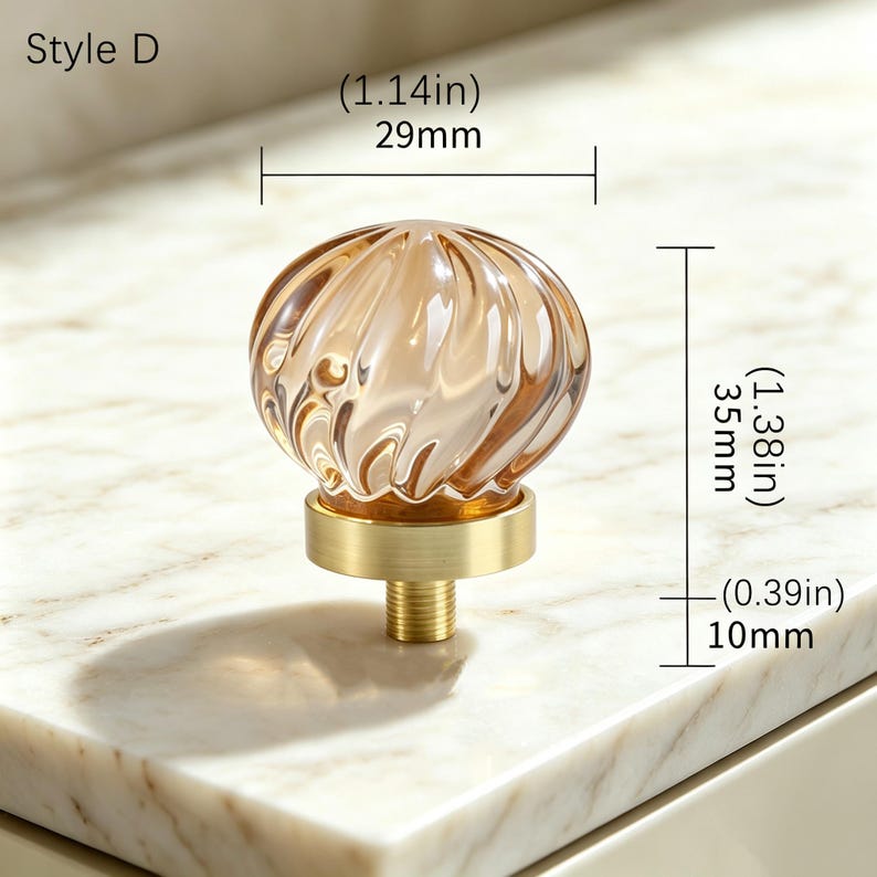 May include: Style D cabinet knob featuring a round, amber-colored glass top with a swirling ribbed texture and a brushed gold metal base. The knob measures 1.14 inches in width, 1.38 inches in total height, and has a 0.39 inch threaded post.