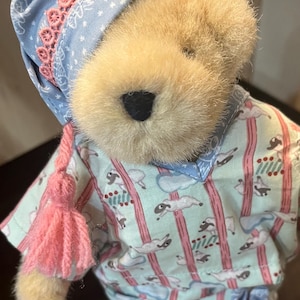 May include: A plush teddy bear wearing a blue and pink sleep cap and pajamas with a cloud and sheep print. The bear has a pink tassel on its cap.